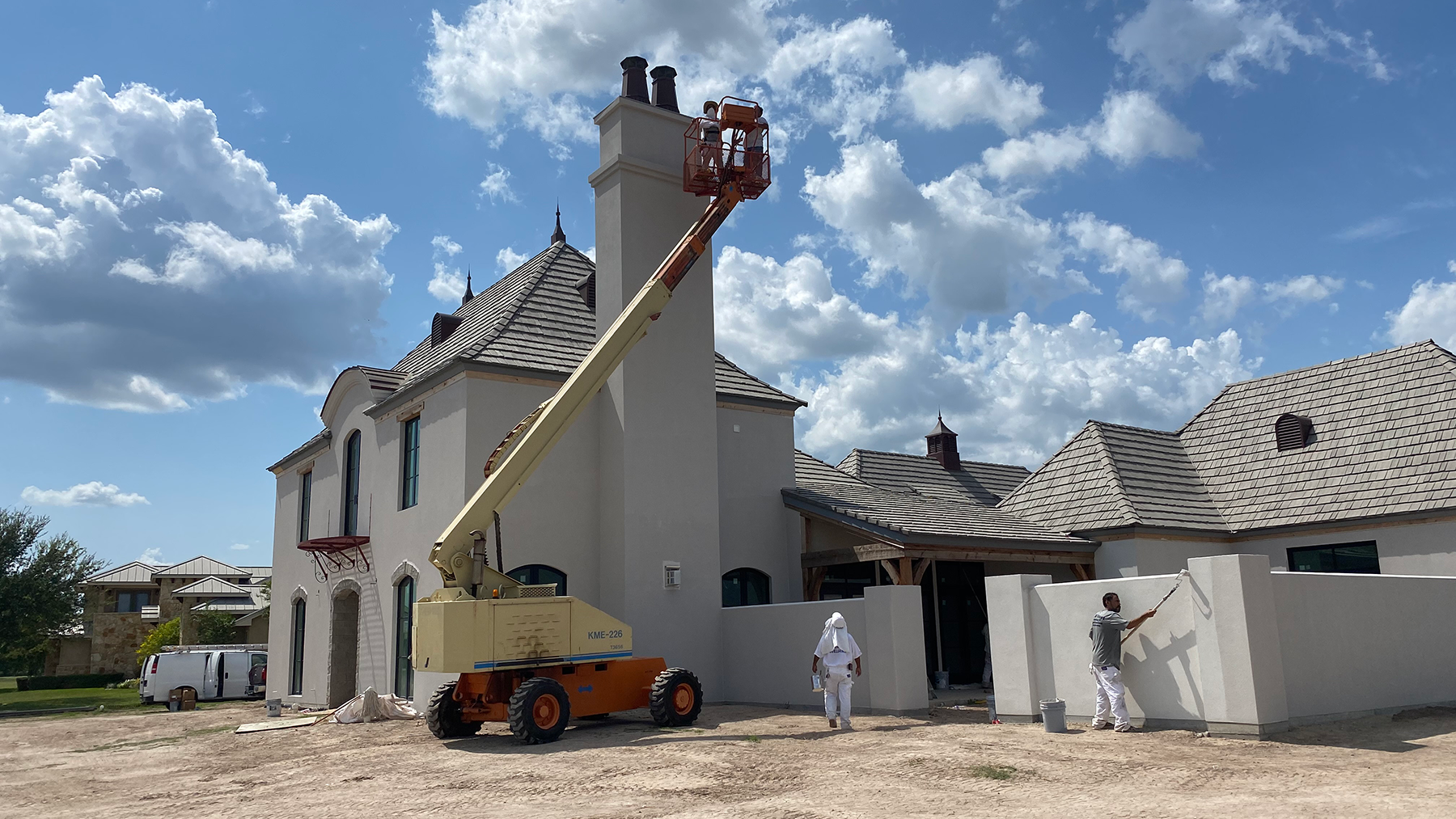 Construction workers using a boom lift to paint a large residential home with a chimney and sloped roof under a cloudy sky