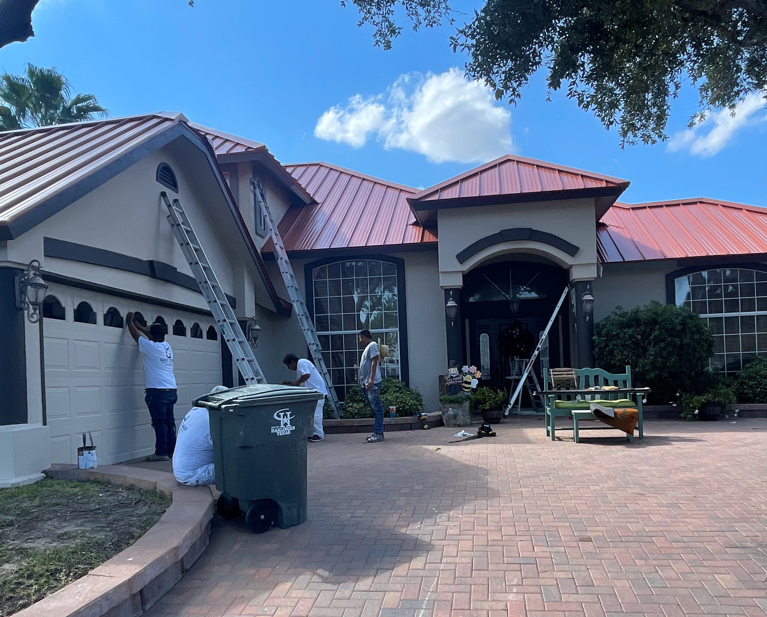 A team of workers painting the exterior of a large, single-story house with a red metal roof. Ladders are set up against the house, and various supplies are placed around the front, including a trash bin and a bench.