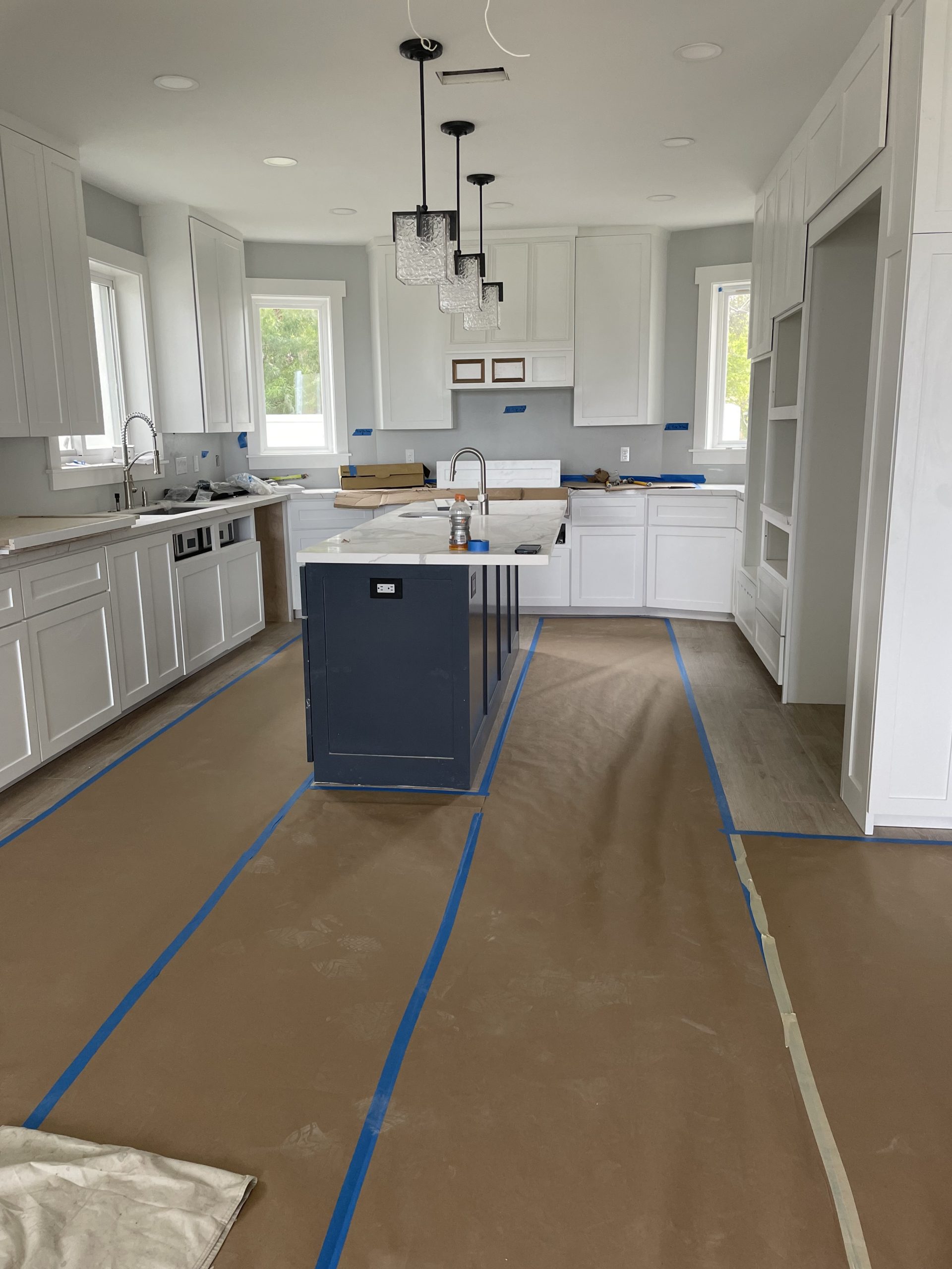 A kitchen with white cabinets and an island. The floor is covered with brown protective paper secured with blue tape, and there are pendant lights above the island